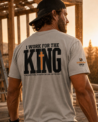 A man wearing a light gray crewneck t-shirt with a large black graphic on the back reading 'I WORK FOR THE KING' with a cross over the letter 'I'. The shirt also features a small 'Kings of NY Crew' logo on the left sleeve.