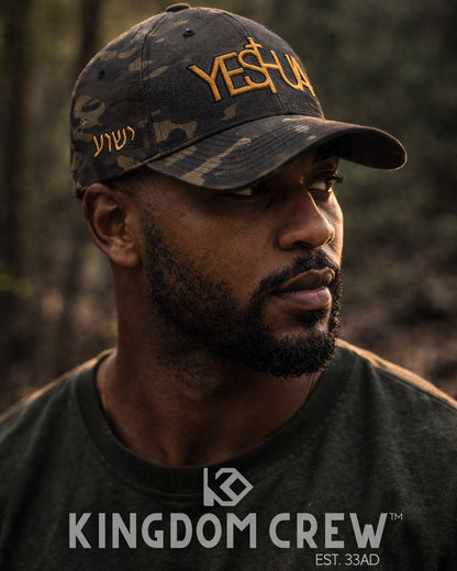 Man wearing a camouflage cap with 'YESHUA' and a black shirt with 'KINGDOM CREW' branding in a forest setting.