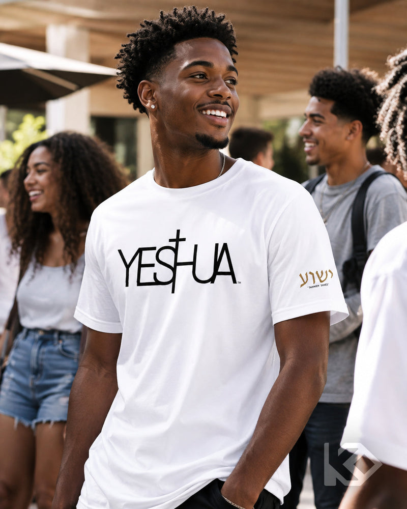 A smiling young man wearing a white graphic T-shirt with the word 'YES' in black and a cross symbol, plus gold script on the sleeve, photographed outdoors with other people blurred in the background.