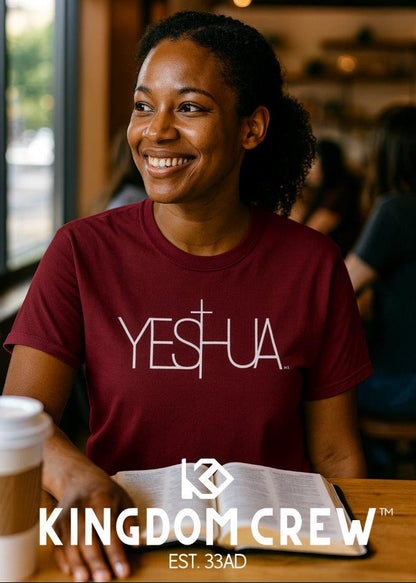 Woman wearing a maroon 'YESHUA' shirt sitting at a table with a coffee cup and book, with 'Kingdom Crew' branding.
