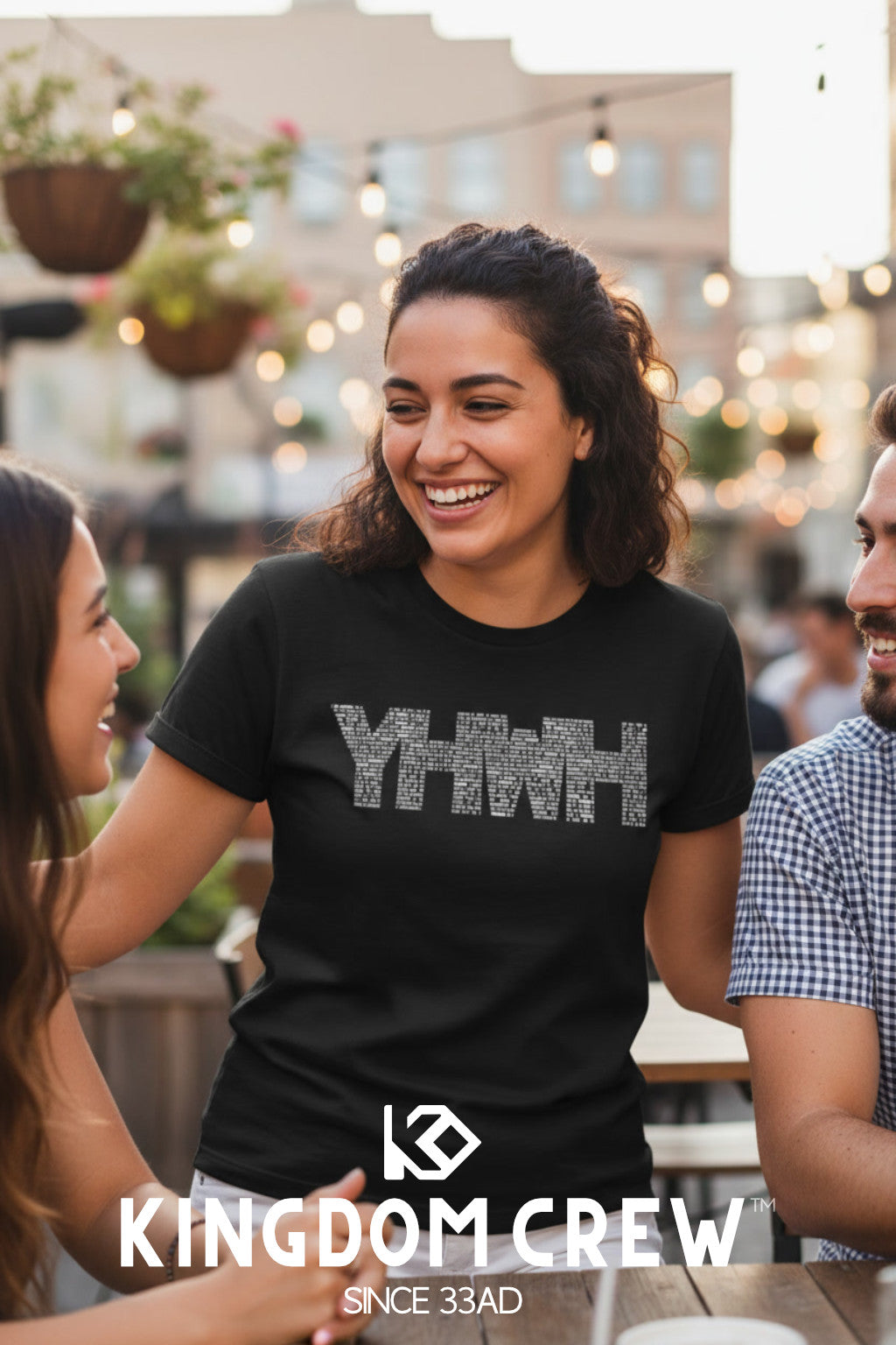 Woman wearing a black t-shirt with 'YHWH' printed on it, sitting outdoors with friends.